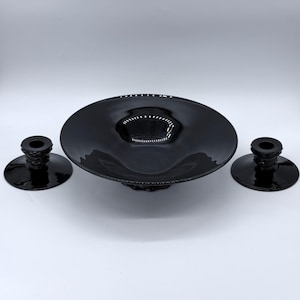 Puede incluir: Un juego de centro de mesa de vidrio negro, que incluye un cuenco grande y poco profundo y dos candelabros a juego. El cuenco tiene un borde ancho y plano y un centro ligeramente elevado. Los candelabros tienen una base redonda y una parte superior cilíndrica.