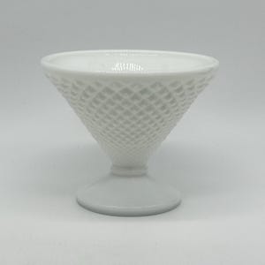 Puede incluir: Un plato de postre blanco de estilo vintage. El plato de vidrio tiene forma de cono con patrón de diamantes, un tallo pequeño y una base redonda. El borde es liso y el plato es de vidrio blanco opaco.
