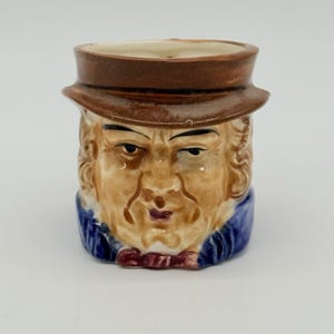 Puede incluir: Una taza de cerámica con forma de cabeza de hombre, con sombrero marrón y rostro bronceado detallado. La taza tiene una chaqueta azul y una base pequeña. La abertura es ancha.