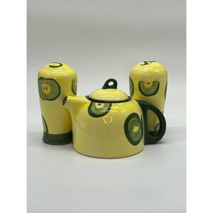 May include: A yellow ceramic teapot with a black handle and lid, accompanied by two matching cylindrical containers. Each piece is decorated with green apple slice designs. The set is designed for serving tea.