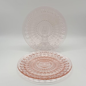 May include: Two pink glass plates with detailed embossed patterns. The plates feature a circular design with a central medallion and radiating lines. One plate is displayed on a clear stand, highlighting the texture and design. Ideal for serving, these plates are a decorative addition.
