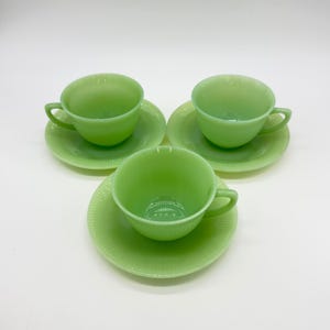 Puede incluir: Tres tazas de té de vidrio translúcido de color verde claro con sus platos a juego. Cada taza presenta una forma de cuenco redondeado con un asa lateral y una textura acanalada. Los platos tienen un patrón acanalado que irradia desde el centro.
