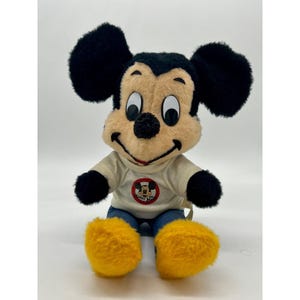 May include: A plush Mickey Mouse doll with black ears, black arms, and yellow feet. Mickey wears a white shirt with a circular graphic and blue jeans. The doll has a tan face with a black nose and a smiling red mouth.