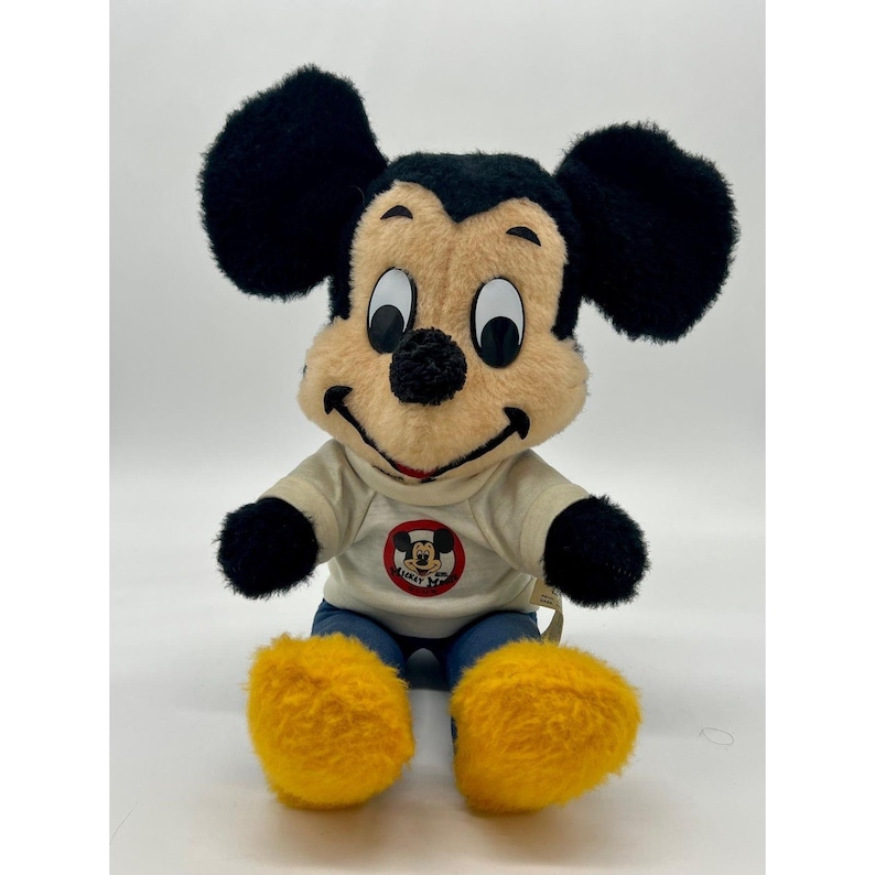 May include: A plush Mickey Mouse doll with black ears, black arms, and yellow feet. Mickey wears a white shirt with a circular graphic and blue jeans. The doll has a tan face with a black nose and a smiling red mouth.