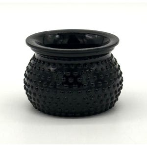 Könnte beinhalten: Eine kleine, runde, schwarze Glasschale mit einer strukturierten, noppenartigen Oberfläche. Die Schale hat einen breiten, glatten Rand und einen leicht bauchigen Körper. Die dunkle Farbe und die einzigartige Textur verleihen ihr ein Vintage- oder Antik-Aussehen.