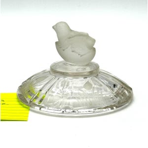 May include: A clear glass perfume bottle with a frosted glass chick emerging from an egg-shaped stopper. The bottle has a round, ridged base with etched floral designs. The overall design is delicate and elegant.