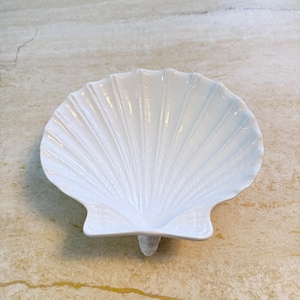 May include: A white, seashell-shaped dish with a textured surface. The dish has a scalloped edge and a small, rounded base. The dish is made of ceramic and is likely used for serving or display.
