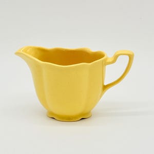 May include: A bright yellow ceramic creamer pitcher featuring a scalloped rim, a curved handle, and a footed base. The pitcher has a smooth, solid-colored finish with a distinct pouring spout and a fluted body design that adds texture to the piece.