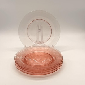 Può includere: Un set di piatti in vetro rosa con un motivo floreale. I piatti sono impilati su un supporto trasparente, con un piatto dietro la pila. I piatti hanno un'estetica delicata e vintage, adatti per servire.