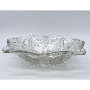 May include: Clear glass serving bowl with a starburst and geometric pattern. The bowl has a scalloped edge and is designed with a series of repeating shapes. The bowl is likely used for serving food or as a decorative piece.
