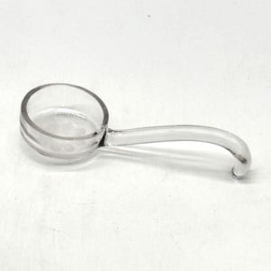 Könnte beinhalten: Eine kleine Schöpfkelle aus klarem Glas mit einer kreisförmigen Schale und einem gebogenen Griff. Das Utensil besteht aus transparentem Glas und weist eine glatte, polierte Oberfläche auf, die sich von der runden Schale bis zum Ende erstreckt.