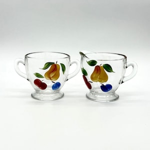 May include: A clear glass sugar bowl and creamer set. Each piece is decorated with hand-painted yellow pears, red cherries, blue berries, and green leaves. The sugar bowl has two handles.