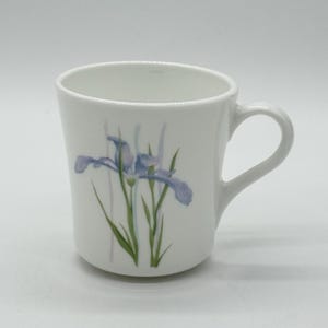 May include: White ceramic mug with a curved handle. The mug features a painted design of a blue iris flower with green leaves and stems. The design is centered on the front of the mug.