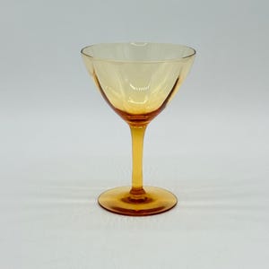Puede incluir: Una copa de cristal de color ámbar translúcido con un cuenco ancho y poco profundo, un tallo delgado y una base circular. El cristal presenta un acabado liso y pulido con un tono naranja dorado uniforme en toda la estructura del recipiente.