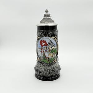 May include: A black ceramic beer stein with a silver lid. The stein features a colourful image of the Swiss flag, people in traditional clothing, and a mountain scene. The word "Switzerland" is also visible.