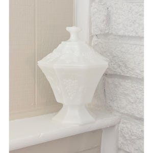Puede incluir: Un cuenco blanco con tapa y base de pedestal. El cuenco tiene una forma geométrica con diseños en relieve de uvas y hojas. La tapa tiene un pequeño pomo para facilitar la extracción. El cuenco es de vidrio opaco.