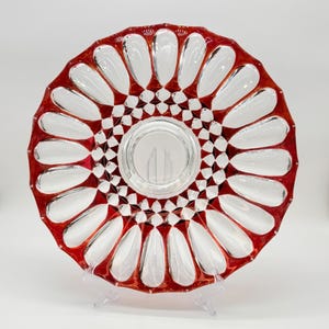 May include: A circular glass plate featuring a vibrant red rim and a clear center. The design includes a series of elongated, petal-shaped indentations radiating from the middle, surrounded by a geometric pattern of small, clear diamond-shaped facets.
