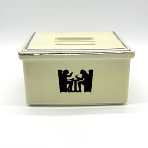 Peut inclure: Un beurrier rectangulaire en céramique de couleur crème avec un couvercle assorti. L'avant de la base présente une silhouette noire représentant deux personnages assis à une petite table. Le couvercle a une poignée rectangulaire.