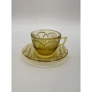 May include: A vintage amber-colored teacup and saucer set. The cup and saucer are made of glass and feature a decorative embossed pattern. The cup has a handle and the saucer is round. The set is likely from the mid-20th century.