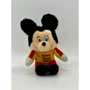 May include: A plush Mickey Mouse toy with black ears, a tan face, and a yellow shirt with red stripes and the text "Mickey Mouse Power." The toy has white gloves and red shorts. The toy is sitting on a white surface.
