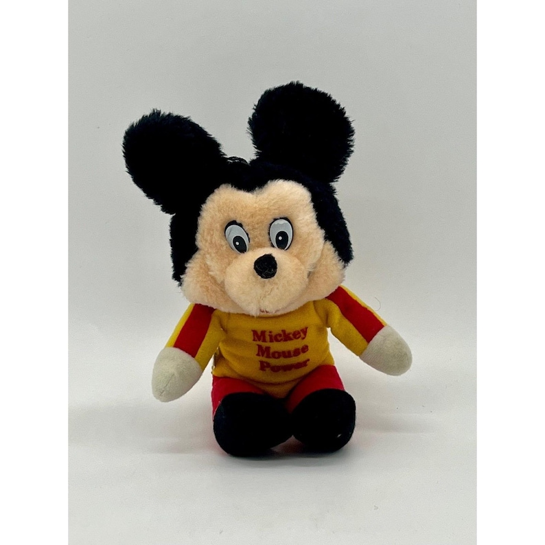 May include: A plush Mickey Mouse toy with black ears, a tan face, and a yellow shirt with red stripes and the text "Mickey Mouse Power." The toy has white gloves and red shorts. The toy is sitting on a white surface.
