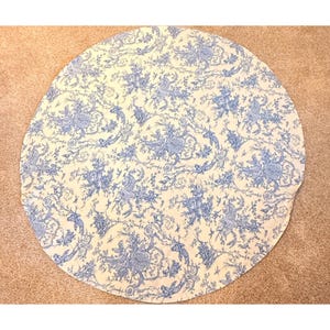 May include: A round, cream-colored tablecloth with a blue floral toile pattern. The design features detailed illustrations of flowers, birds, and foliage. The tablecloth is made of a soft fabric and is suitable for a dining table.