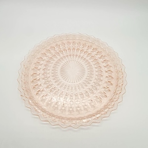 May include: A light pink glass serving plate with a detailed geometric pattern. The plate has a scalloped edge and a central design radiating outwards. The plate is likely vintage and suitable for serving desserts or appetizers.