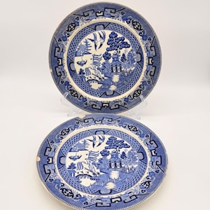 May include: Two blue and white ceramic plates with a Willow pattern design. The plates feature a central scene with a pagoda, trees, and figures, surrounded by a decorative border. The plates are displayed on a clear stand against a white background.