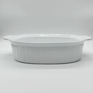 May include: A white, oval-shaped ceramic baking dish with a ribbed exterior and two handles. The dish is empty and sits on a white surface. The dish is designed for baking and serving.