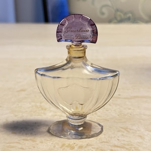 Puede incluir: Un frasco de perfume de vidrio transparente con un tapón morado. El tapón es arqueado y tiene las palabras "Guerlain Paris" impresas. El frasco tiene una forma única con una base ancha y un cuello estrecho.
