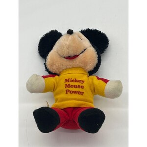 May include: A plush Mickey Mouse toy with black ears, a tan face, and a red mouth. Mickey wears a yellow shirt with "Mickey Mouse Power" printed on it, red pants, and white gloves. The toy is sitting.