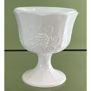 Puede incluir: Una copa blanca de estilo vintage con base de pedestal. El vidrio presenta un diseño texturizado de uvas y hojas en el cuenco. La copa tiene forma hexagonal y está hecha de vidrio blanco opaco.