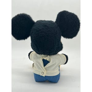 May include: A vintage Mickey Mouse plush toy, viewed from the back. The plush features large black ears, a black head, a white shirt, and blue denim pants. The shirt has a button closure. The toy is a classic Disney collectible.