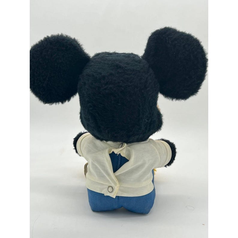 May include: A vintage Mickey Mouse plush toy, viewed from the back. The plush features large black ears, a black head, a white shirt, and blue denim pants. The shirt has a button closure. The toy is a classic Disney collectible.