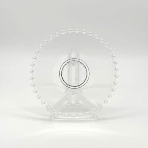 Op de afbeelding: Een heldere, decoratieve plaat met een kralenrand, gepresenteerd op een heldere plastic standaard. De plaat heeft een centraal cirkelvormig ontwerp. De standaard heeft een driepotige basis. Het ontwerp is minimalistisch en elegant. Diameter: ongeveer 20 cm.