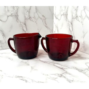 May include: Two ruby red glass serving pieces. One is a creamer with a spout and handle, and the other is a handled cup. Both pieces have a simple, classic design and are made of transparent glass.