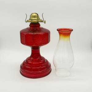 Op de afbeelding: Een olielampvoet van rood glas met een getextureerd, gefacetteerd oppervlak en een messing brandermechanisme, vergezeld van een losse glazen lampenkap met een oranje-rode kleurverloop aan de bovenrand en een gedetailleerde kralenrand.