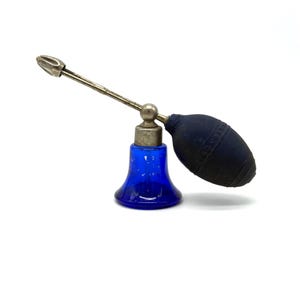 Puede incluir: Un frasco de perfume azul cobalto vintage con un atomizador plateado. El frasco tiene una base bulbosa y un tubo metálico largo y delgado con una punta decorativa. La pera del atomizador es azul oscuro.