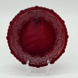 May include: A ruby red glass plate with a scalloped edge and embossed floral and geometric patterns. The plate has a textured center and is displayed on a clear plastic stand. This vintage-style plate is suitable for serving food or as a decorative piece.