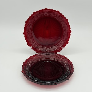 May include: Two ruby red glass plates with a scalloped edge and ornate embossed design. The plates have a central sunburst pattern and are displayed on a white background. The plates are likely for decorative or serving purposes.