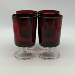May include: Four ruby red glass goblets with a short stem and a round base. The glasses have a cylindrical shape and are arranged in a square formation. The background is plain white.