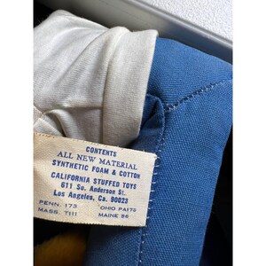 May include: Close-up of a vintage stuffed toy with a blue fabric body and white fabric collar. A tag reads "CONTENTS ALL NEW MATERIAL SYNTHETIC FOAM & COTTON CALIFORNIA STUFFED TOYS 611 So. Anderson St. Los Angeles, Ca. 90023".