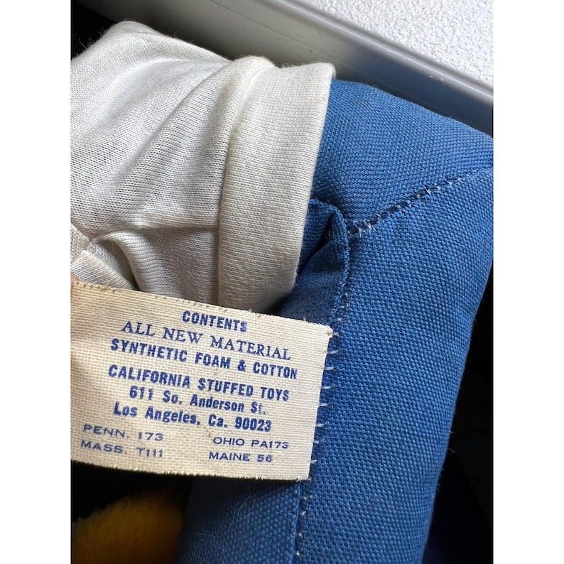 May include: Close-up of a vintage stuffed toy with a blue fabric body and white fabric collar. A tag reads "CONTENTS ALL NEW MATERIAL SYNTHETIC FOAM & COTTON CALIFORNIA STUFFED TOYS 611 So. Anderson St. Los Angeles, Ca. 90023".