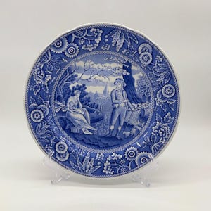 May include: A blue and white decorative plate with a pastoral scene in the center. The plate features a scalloped edge and floral border. The central image depicts figures near a tree and a building in the background.