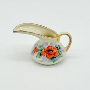May include: A small ceramic creamer jug with a cream-coloured base, featuring a hand-painted orange poppy flower with green leaves and light purple accents. The rim and the small handle are trimmed with a thin line of metallic gold paint.