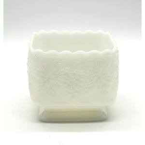 Puede incluir: Un cuenco cuadrado de vidrio blanco vintage con borde festoneado. Los lados presentan un detallado patrón floral en relieve. El cuenco se asienta sobre una pequeña base rectangular. El diseño general es delicado y ornamentado.