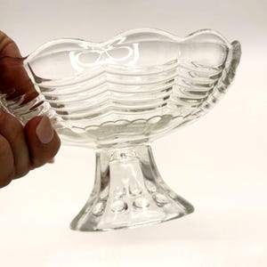 Puede incluir: Un cuenco de postre de cristal transparente y vintage, con borde festoneado y base de pedestal. El cuenco tiene un diseño ondulado y texturizado. La base presenta pequeñas hendiduras redondeadas. El cuenco es sostenido por una mano.