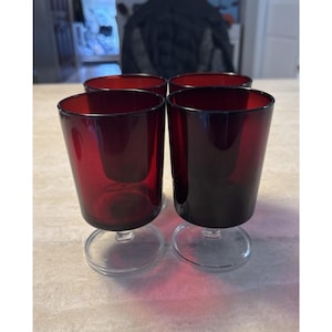 May include: Four red-tinted glass goblets with clear, circular bases. The glasses have a cylindrical shape and are arranged closely together. The goblets are likely for serving beverages and have a vintage aesthetic.