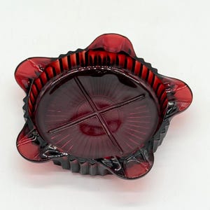 Peut inclure: Un cendrier vintage en verre rouge rubis. Le cendrier présente un motif en étoile au centre et un bord festonné. Le bord comporte une série de nervures verticales. Le cendrier est d'une couleur rouge profond et transparente.