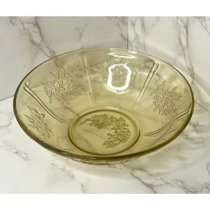 May include: A clear, yellow-tinted glass bowl with floral embossed designs. The bowl has a wide, shallow shape with a rounded base and a decorative floral pattern in the center. The sides feature vertical lines and additional floral motifs.
